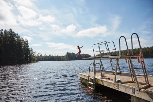 Barn hoppar från en badbrygga ut i en skogssjö en solig sommardag