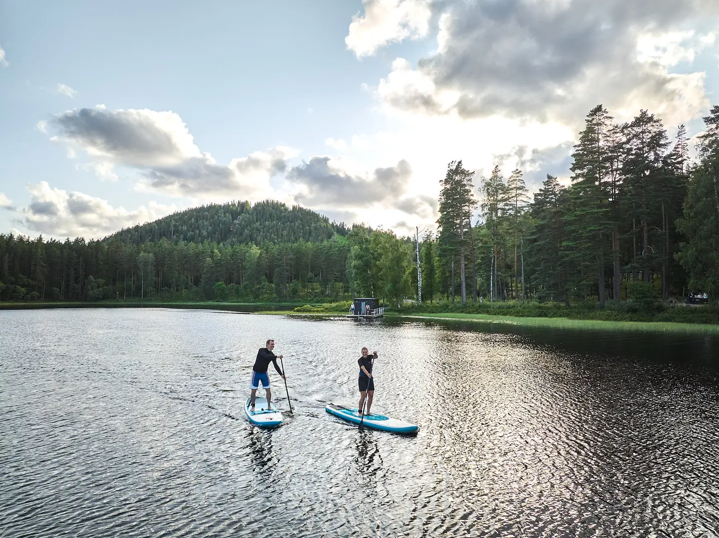 Två personer paddlar SUP på en blank sjö omgiven av skog och en rundad bergstopp